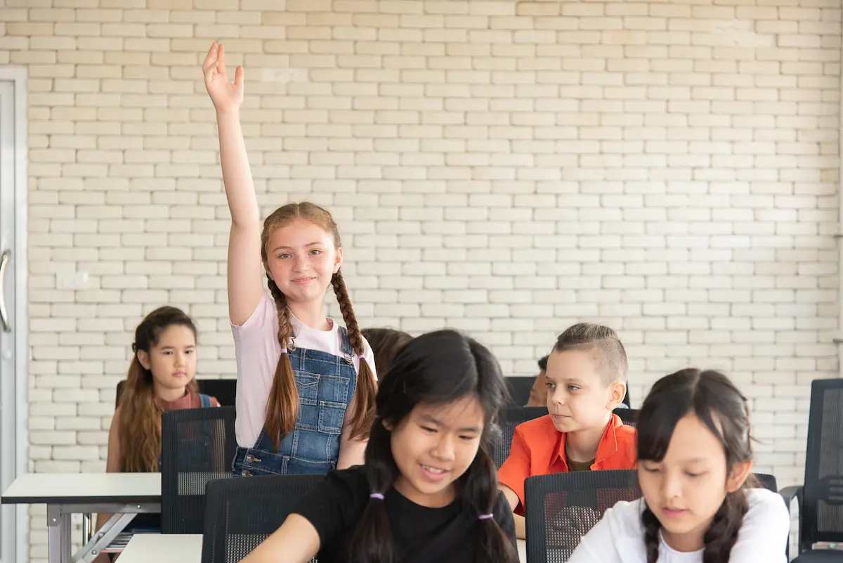 A female student stands in a diverse classroom and raises her hand confidently, demonstrating an inclusive classroom. 